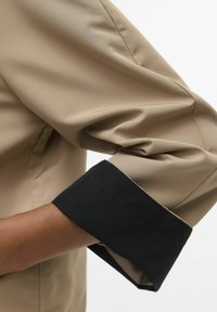 Beige garo piedurknēm krekls ar melnu aproci. Audums izskatās gluds un strukturēts, ar vieglu spīdumu. Aproce ir kārtīgi izšūta, kontrastējot ar krekla ķermeni.