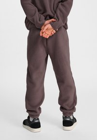 Fleece-Jogginghosen in Dunkelbraun mit elastischen Bündchen und lockerer Passform, kombiniert mit schwarz-weißen Sneakers. Die Textur wirkt weich.