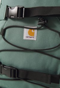 Groene rugzak van duurzaam materiaal, met zwarte riemen, een gesp en een Carhartt-logo met een oranje ontwerp.