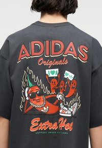 Πίσω μέρος μαύρου πουκάμισου με κόκκινο κείμενο "ADIDAS Originals", καρτούν πιπεριές τσίλι με γυαλιά ηλίου και αθλητικά παπούτσια, φλόγες και το σύνθημα "Extra Hot".