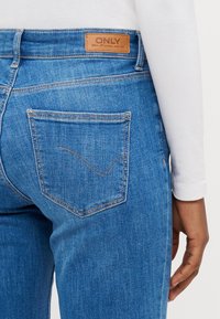 Modré džíny z denimu s hladkou texturou, se hnědou koženou štítkem na zadní straně a dvěma zadními kapsami s zakřiveným švem.