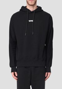 Uomo che indossa un hoodie nero con lacci e una piccola scritta bianca sul petto, abbinato a pantaloni neri, su uno sfondo neutro.