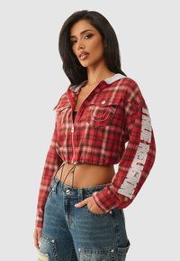 Vrouw in een cropped rood geruit jack met ritssluiting en de tekst "TRUE RELIGION" op de mouw, met blauwe distressed jeans tegen een effen achtergrond.