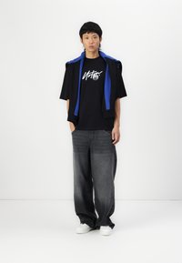 Black T-shirt with white graphic and silver accent, paired with loose gray jeans and white sneakers, layered with a blue and black jacket.