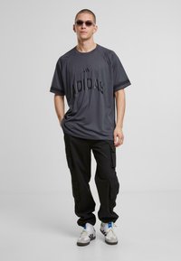 Hallitusülikond, millel on sissepressitud "ADIDAS" logo, mustade cargo-pükstega ja valgete kingadega, millel on sinised triibud. Kandes prille.