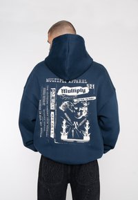 Sweat à capuche oversize bleu marine avec un imprimé graphique dans le dos, comportant du texte en gras et des illustrations. Fabriqué en tissu doux, il a une grande capuche.