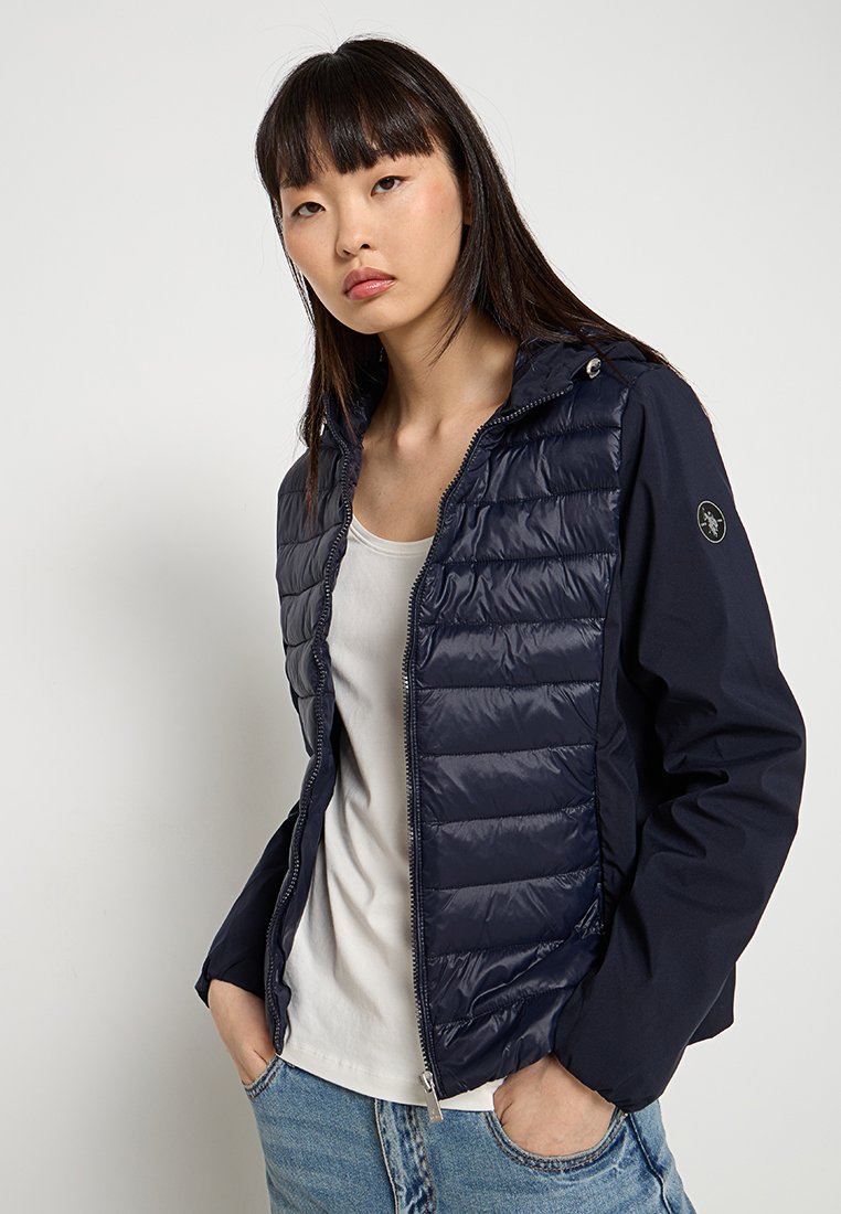 Jonge vrouw met steil, donker haar draagt een marineblauwe gewatteerde jas over een wit shirt en blauwe jeans, staand tegen een lichte achtergrond.