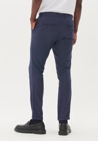 Pantalones de vestir en azul marino con una textura sutil y un corte ajustado, que presentan puños doblados y bolsillos traseros con cierres de botón. Incluidos unos zapatos de cuero negros.