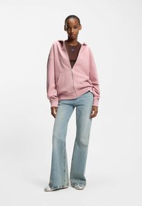 Light pink zip-up hoodie with a large front pocket, relaxed fit, and ribbed cuffs, worn over a brown top and paired with flared blue jeans.