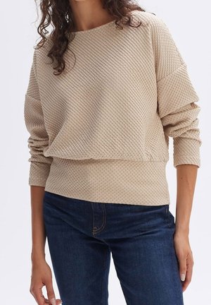 Frau trägt beigen, strukturierten Langarmpullover mit überschnittenen Schultern und dunkelblaue Jeans, steht vor einfachem Hintergrund.