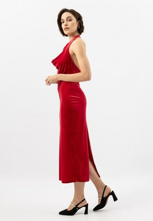 Abito halter in raso rosso con spacco laterale, silhouette aderente e texture liscia. Indossato con décolleté nere con tacco a cui si aggiungono decorazioni sul fascione.