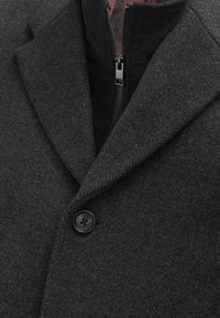 Abrigo de lana gris oscuro con solapa, que presenta un interior negro con cremallera y un único botón negro en la parte delantera. Primer plano de la tela texturizada.