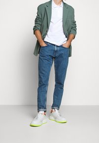 Blazer vert porté sur un polo blanc, jean bleu et baskets blanches sportives avec semelles vert fluo et accents orange.