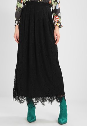 Czarna midi spódnica koronkowa z falowanym brzegiem, zestawiona z turkusowymi satynowymi botkami za kostkę i kwiecistą bluzką z długim rękawem, modelka stoi.