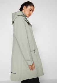 Cappotto impermeabile grigio chiaro con cappuccio, tasca laterale con cerniera e inserti riflettenti. Presenta un taglio dritto e una texture liscia.