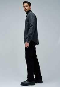 Camisa de mezclilla gris con cuello y botones, combinada con pantalones negros texturizados y zapatos negros tipo slip-on. El modelo está de pie contra un fondo gris.
