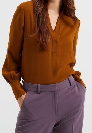 Vrouw die een roestkleurige blouse met lange mouwen draagt, ingestopt in paarse broek met hoge taille, met handen in de zakken, tegen een effen achtergrond.