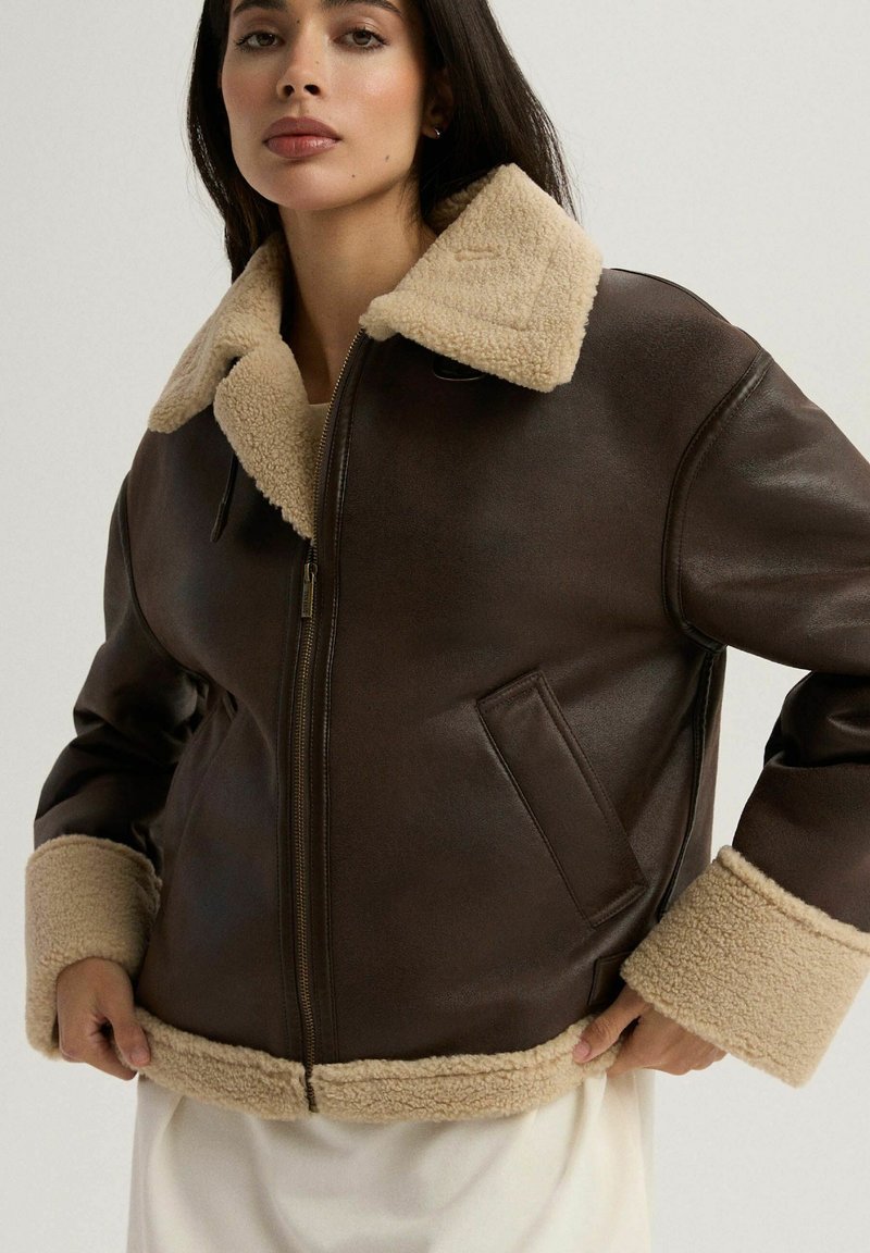 Brązowa skórzana kurtka z beżowym kołnierzem i mankietami z owczej wełny. Posiada zamek błyskawiczny z przodu, kieszenie boczne oraz luźny krój. Miękka struktura.