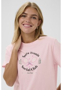 Smaidoša blondīne ar zilām acīm, valkā zeltainas riņķa auss gredzenus un rozā T-kreklu ar krustotiem tenisa raketēm un tenisa bumbiņu grafiku.