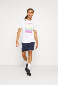 White t-shirt with a colorful geometric graphic, paired with navy shorts. Black athletic shoes and white socks with logo accents visible.