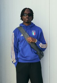 Μπλε φούτερ με κίτρινες ρίγες και σήμα Ιταλίας, μαύρο παντελόνι και μαύρη τσάντα χιαστί. Μοντέλο που φοράει γυαλιά ηλίου, στέκεται μπροστά σε ανοιχτό φόντο.