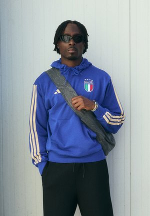 Blaues Hoodie mit gelben Streifen und Italien-Wappen, schwarze Hose und schwarze Umhängetasche. Modell trägt Sonnenbrille und steht vor einem hellen Hintergrund.