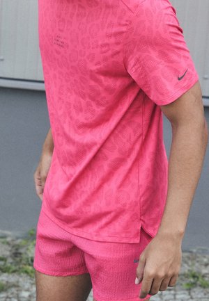 Mężczyzna ubrany w jasnoróżową sportową koszulkę i spodenki z subtelnym zwierzęcym wzorem, stojący na zewnątrz na tle szarej ściany.