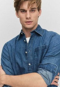 Sininen denim-painikepaita, jossa on etutasku ja käännetyt hihat. Kangas on pehmeän tuntunen ja keskipainoisen muotoilun omaava.