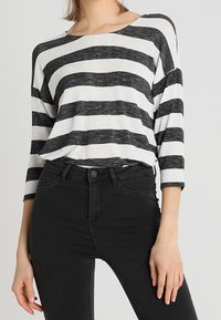 Mustat ja valkoiset raidalliset pitkät hihat, joissa on leveä pääntie ja pehmeä, teksturoitu kangas, yhdistettynä korkeavyötäröisiin mustiin farkkuihin.