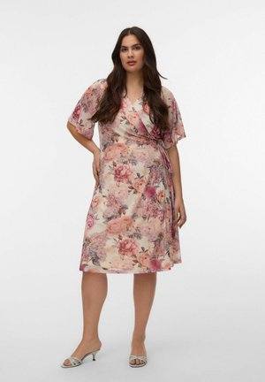 Žena s dlhými tmavými vlasmi v kolenovej dĺžke, v kvetinových zavinovacích šatách a otvorených topánkach na podpätku, stojaca pred jednoduchým bielym pozadím.