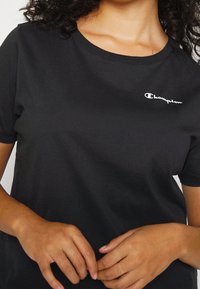 Czarny T-shirt z krótkim rękawem, wykonany z bawełny, z białym haftowanym logo "Champion" na lewej piersi. Gładka faktura.