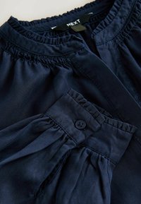 Chemisier bleu marine avec des détails froncés au niveau du col et des poignets, et un unique bouton noir au poignet pour un maintien sécurisé.