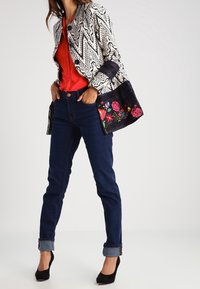 Svart- och vitt mönstrad jacka med blommiga detaljer på ärmarna, tillsammans med en röd topp, mörkblå jeans och svarta högklackade skor.