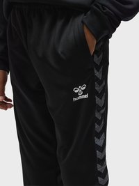 Pantalon de sport noir avec motif chevron latéral et logo hummel, main dans la poche, porté avec un haut noir à manches longues sur fond uni.