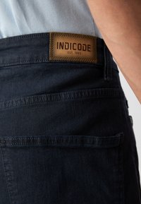 Tamno plave traperice s kožnom etiketom na kojoj piše "INDICODE EST. 1993" blizu stražnjeg pojasnog dijela, s bočnim i stražnjim džepovima.