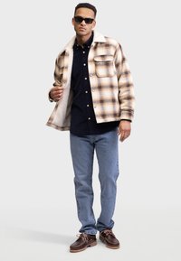 Chaqueta de cuadros en tonos marrón y beige con forro de piel sintética blanca, camisa de botones azul marino, vaqueros azules y zapatos marrones de cordones.