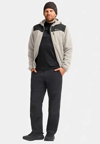 Giacca in pile grigio chiaro con dettagli neri, tasca con cerniera, maglia a manica lunga nera, pantaloni neri e scarpe scure, con cappello lavorato a maglia.