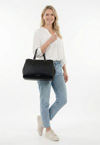 Frau trägt weiße Bluse, hellblaue Jeans und weiße Turnschuhe, hält eine große schwarze Handtasche am Arm und steht vor weißem Hintergrund.