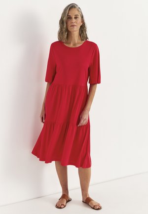 Kvinde iført en rød knælængde kjole med albuelange ærmer og brune flade sandaler stående mod en ensfarvet hvid baggrund.