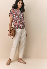 Blouse à motifs multicolores à manches courtes en tissu léger. Associée à un pantalon large couleur crème et des sandales plates.