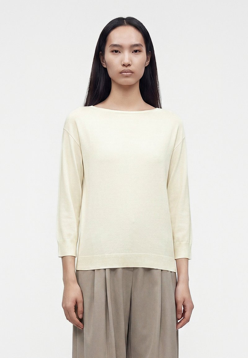 Femme aux longs cheveux foncés portant un haut crème à manches longues et un pantalon beige ample, debout devant un fond blanc uni.