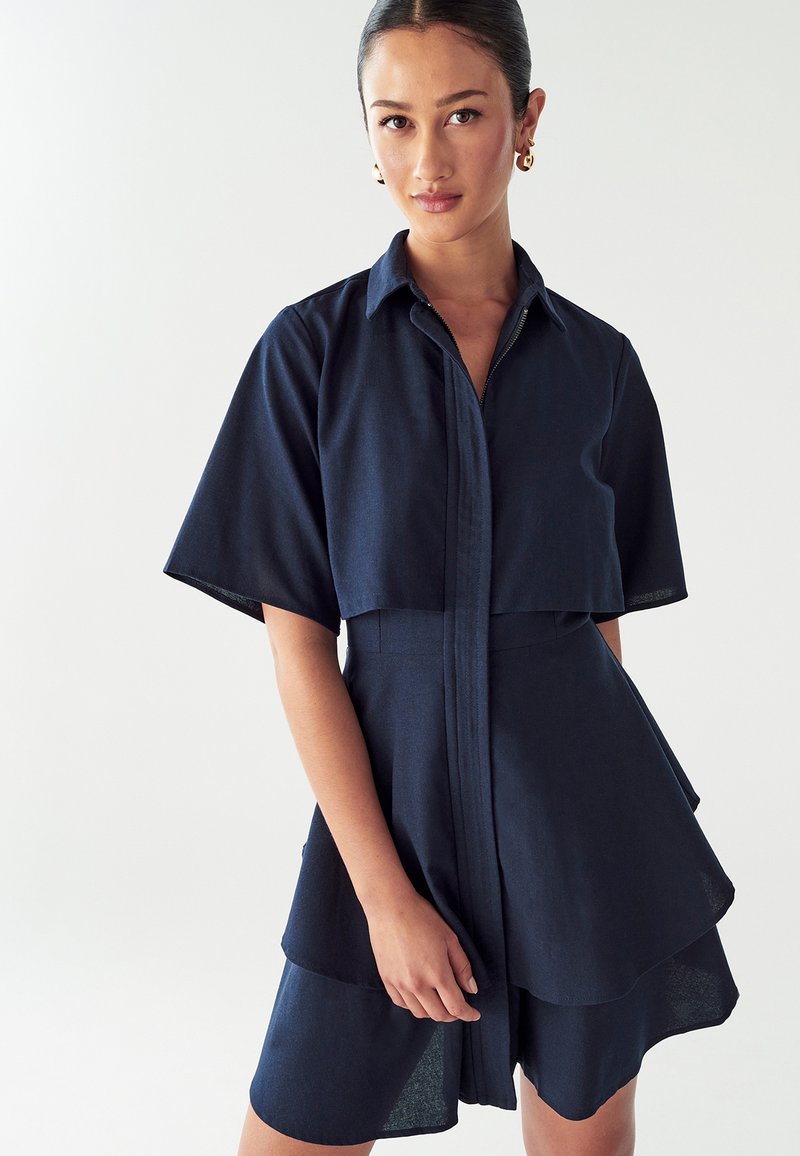 Robe chemise bleu marine à manches courtes, taille cintrée et ourlet à volants superposés. Fabriquée à partir d'un tissu lisse avec un design à boutons sur le devant.