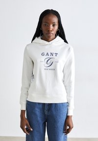 Hvid sweatshirt med hætte, prydet med blåt "GANT" logo og tekst. Kombineret med løse, lyseblå denim jeans. Glat tekstur, afslappet design.