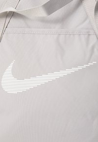 Sac de sport en tissu gris avec un logo Nike à rayures blanches. Texture lisse avec des coutures renforcées et des bretelles robustes.
