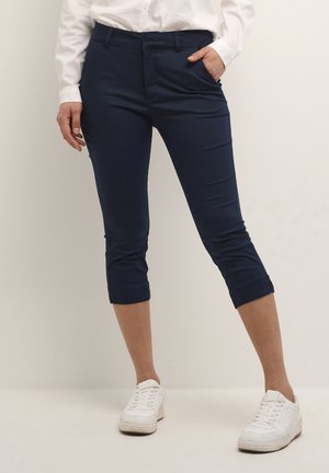 Navy cropped pants made of smooth fabric, featuring a fitted design, front pockets, and turned-up cuffs. Paired with white sneakers.