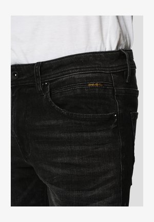 Jean en denim noir avec un design classique à cinq poches, une légère décoloration et un logo doré sur la poche avant. Texture lisse.