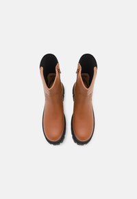 Une paire de bottines en cuir beige avec des panneaux élastiques noirs et des languettes de tirage, vues d'en haut sur un fond blanc.