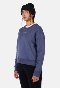 Fioletowy krótki sweatshirt z długimi rękawami, ściągaczami na mankietach i białym logo na klatce piersiowej. Noszony z czarną dzianinową czapką.