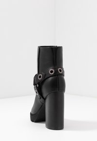Botín negro hasta el tobillo con un tacón grueso, confeccionado en suave cuero sintético. Cuenta con una correa decorativa con acentos metálicos redondos.