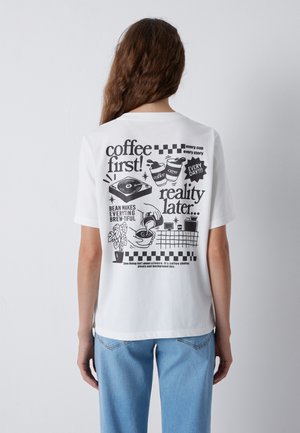 Woman wearing a white t-shirt with black coffee-themed text and illustrations on the back, paired with light blue jeans.
